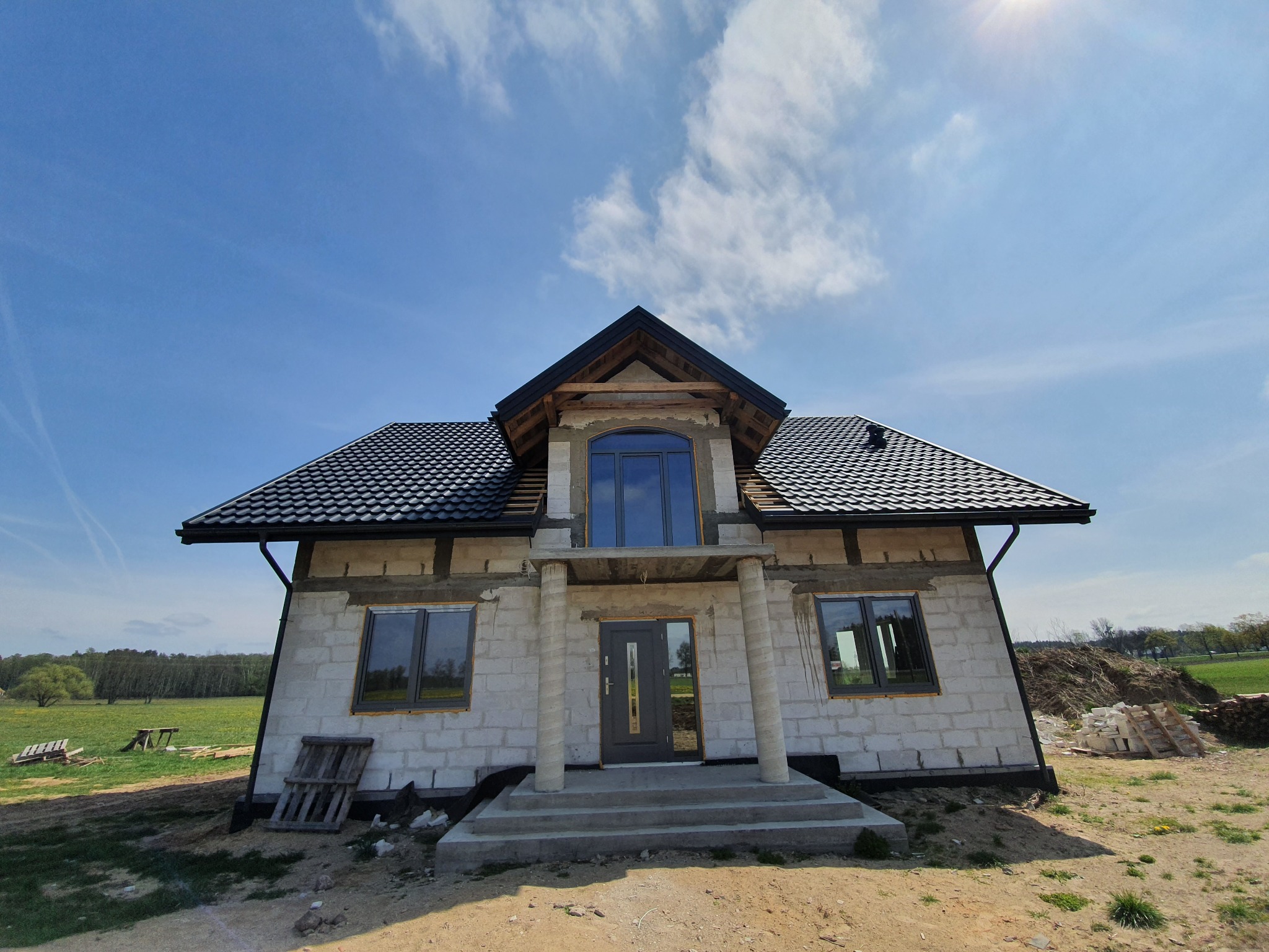 Dom jednorodzinny w trakcie budowy, z widoczną konstrukcją dachu pokrytą czarną dachówką, ściany z białego bloczka, okna i drzwi w kolorze antracytowym, na tle błękitnego nieba.