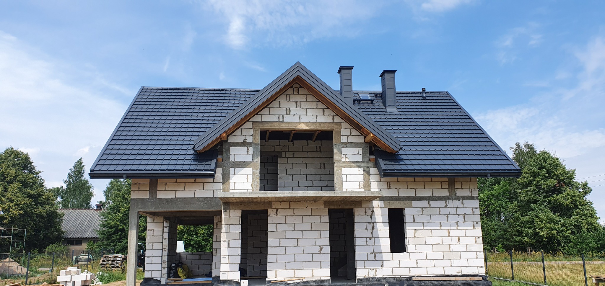 Dom w stanie surowym z pokrytym ciemnoszarą dachówką dachem, widoczne kominy i orynnowanie, na tle błękitnego nieba z chmurami.