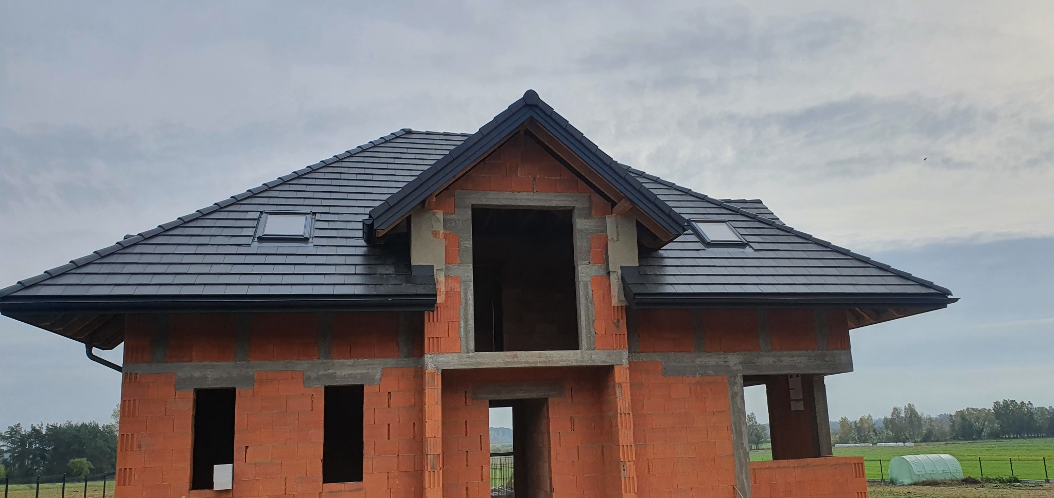 Nowy, ciemny dach z dachówką ceramiczną i oknami połaciowymi na surowym budynku z czerwonej cegły, widoczne rynny i fragment krajobrazu w tle.