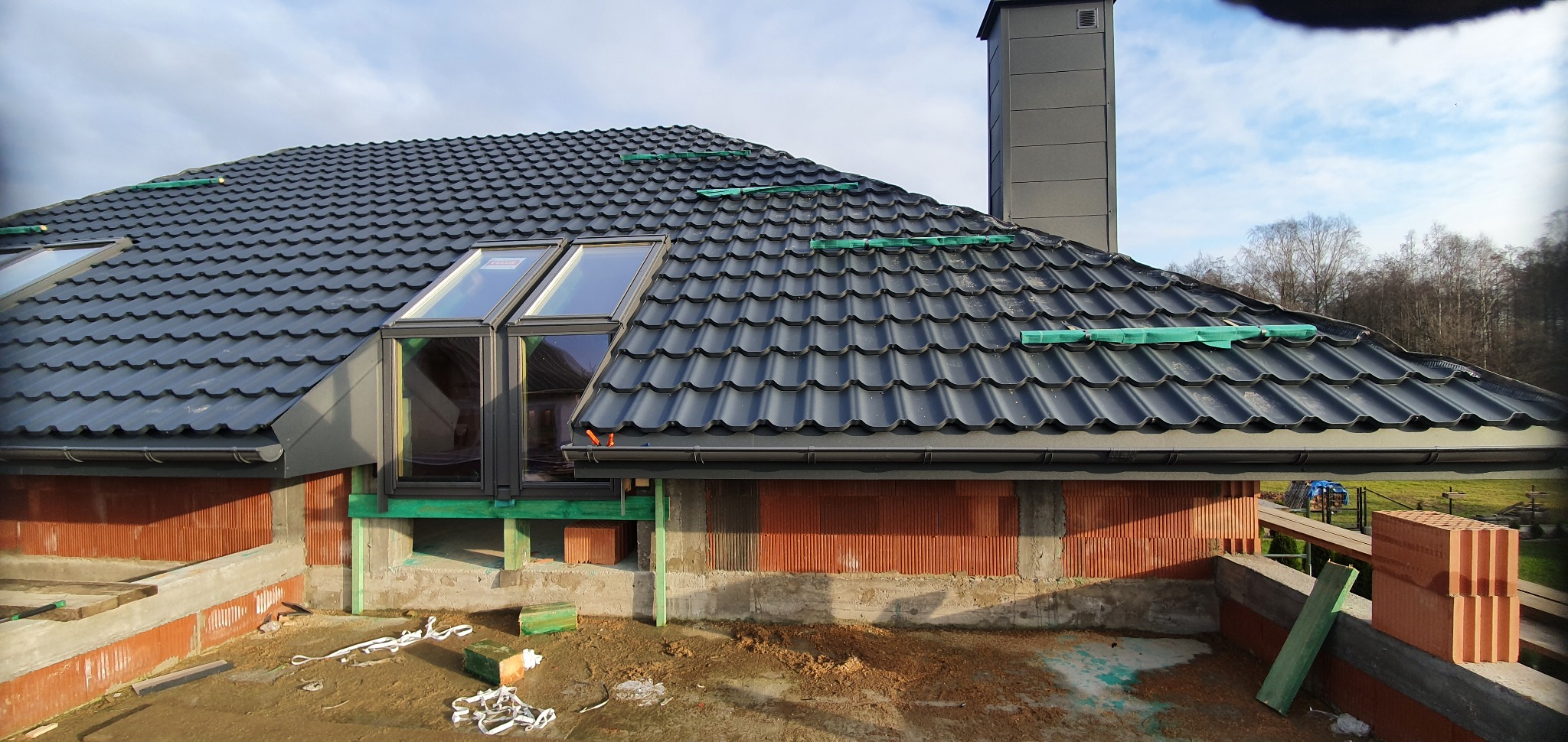 Nowy dach z dachówki ceramicznej w kolorze grafitowym, z oknami połaciowymi i kominem, zabezpieczony zieloną siatką, widok z poziomu robót murarskich