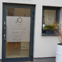 Wejście do Instytutu Kosmetologii Joanna Osuch z logo JO na szkle, podane godziny otwarcia i dane kontaktowe, obok okno z roślinami w doniczce.