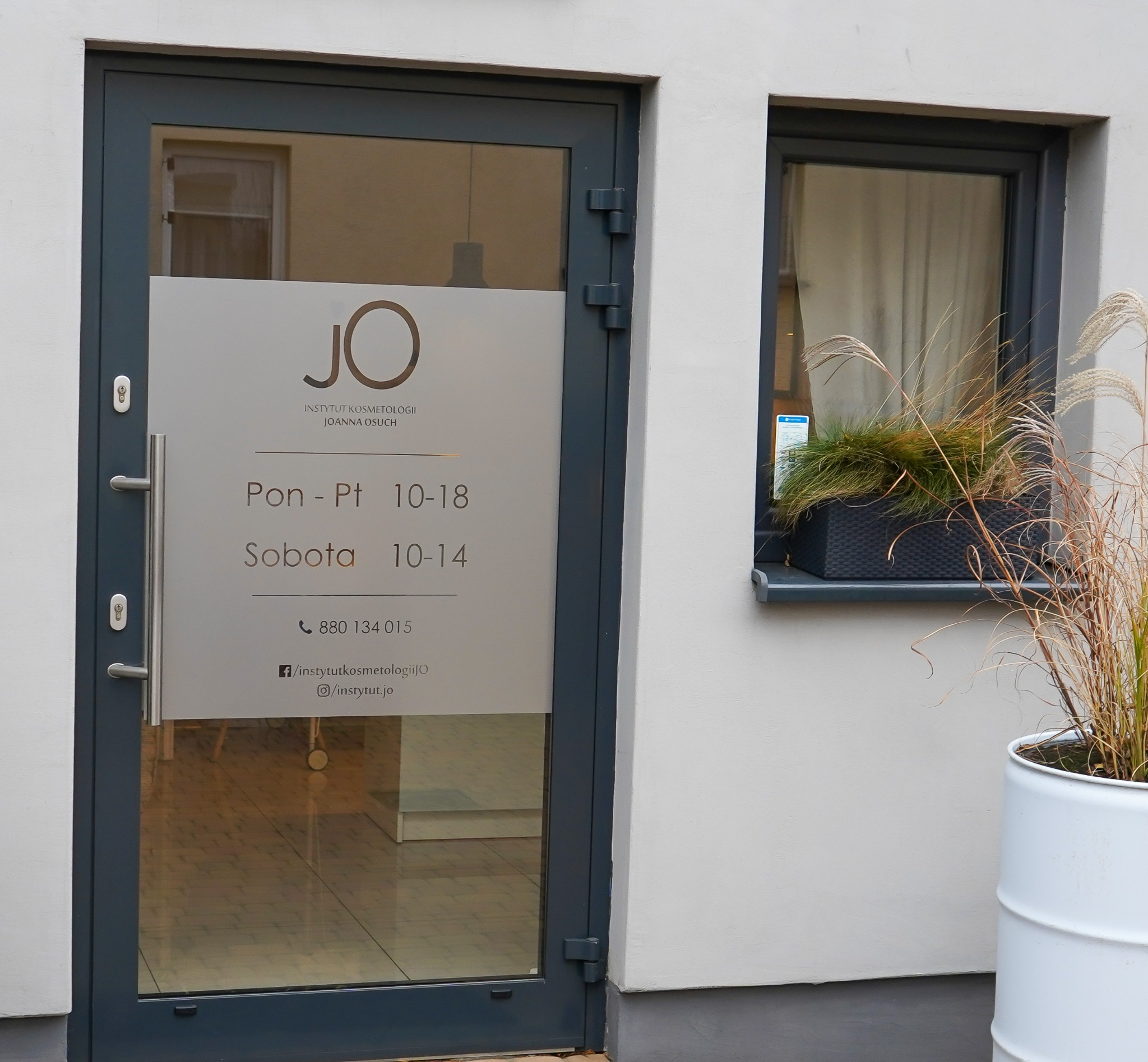Wejście do Instytutu Kosmetologii Joanna Osuch z logo JO na szkle, podane godziny otwarcia i dane kontaktowe, obok okno z roślinami w doniczce.