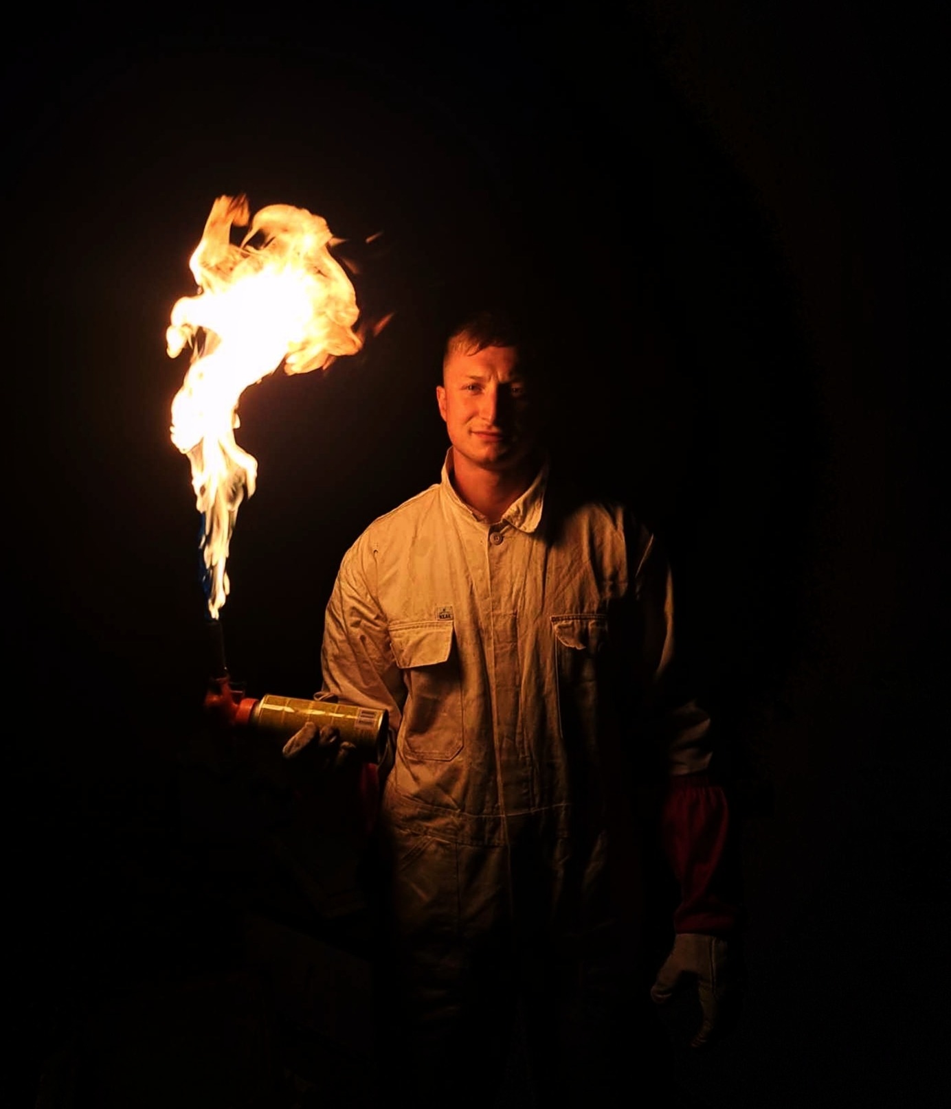 Mężczyzna w kombinezonie ochronnym trzyma palnik gazowy z płomieniem, oświetlony w ciemności, prawdopodobnie w trakcie dezynfekcji uli.