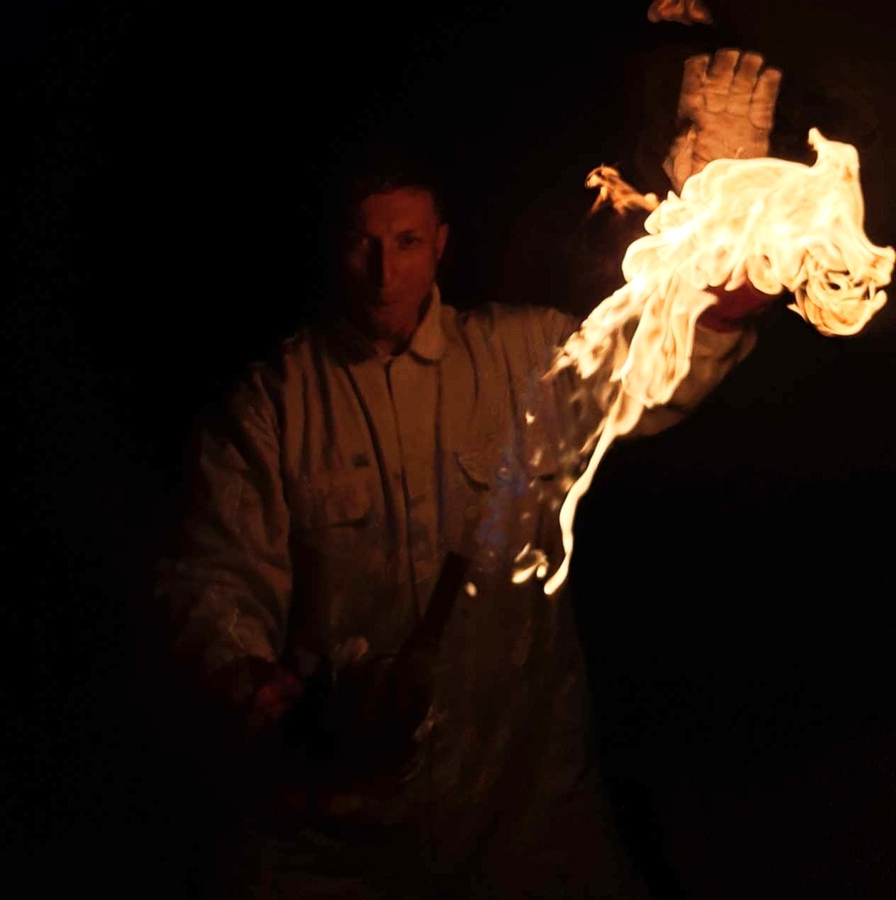 Mężczyzna w kombinezonie trzymający płonącą pochodnię w rękawicy ochronnej, ciemne otoczenie.