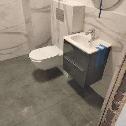 Nowoczesna łazienka z podwieszaną toaletą, szarą szafką pod umywalką i marmurowymi płytkami na ścianach, zabezpieczonymi niebieską taśmą malarską.