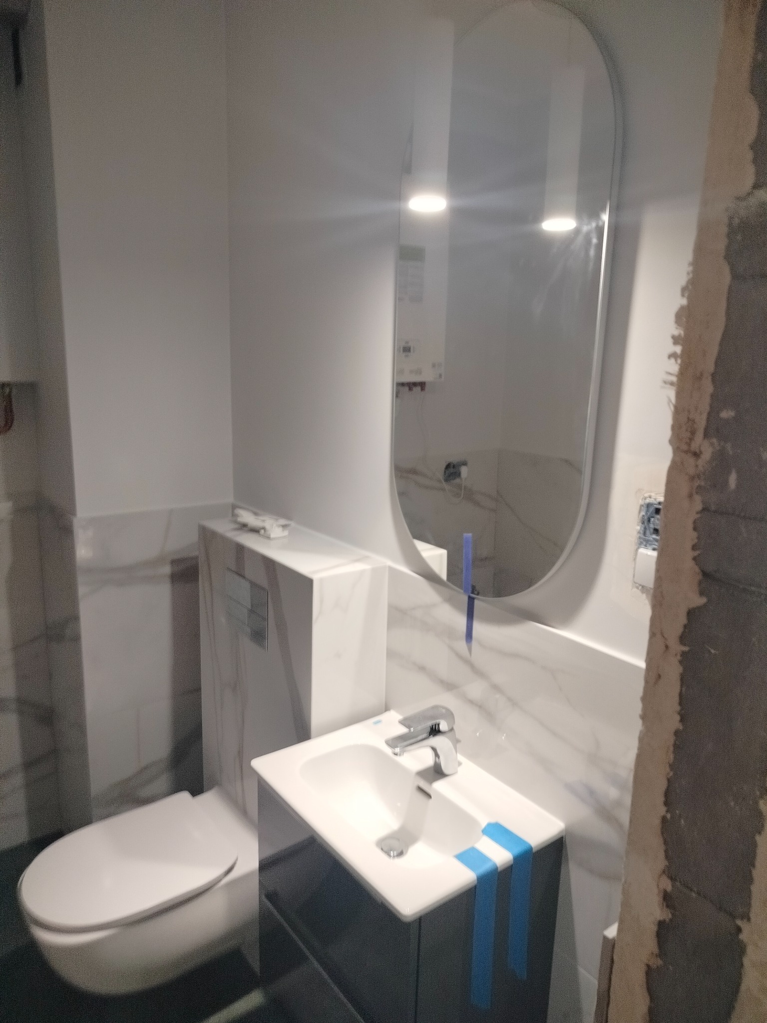 Nowoczesna, biało-szara łazienka w trakcie wykańczania, z owalnym lustrem, umywalką z szafką zabezpieczoną niebieską taśmą i podwieszaną toaletą; widoczne ślady po pracach budowlanych na ścianie.