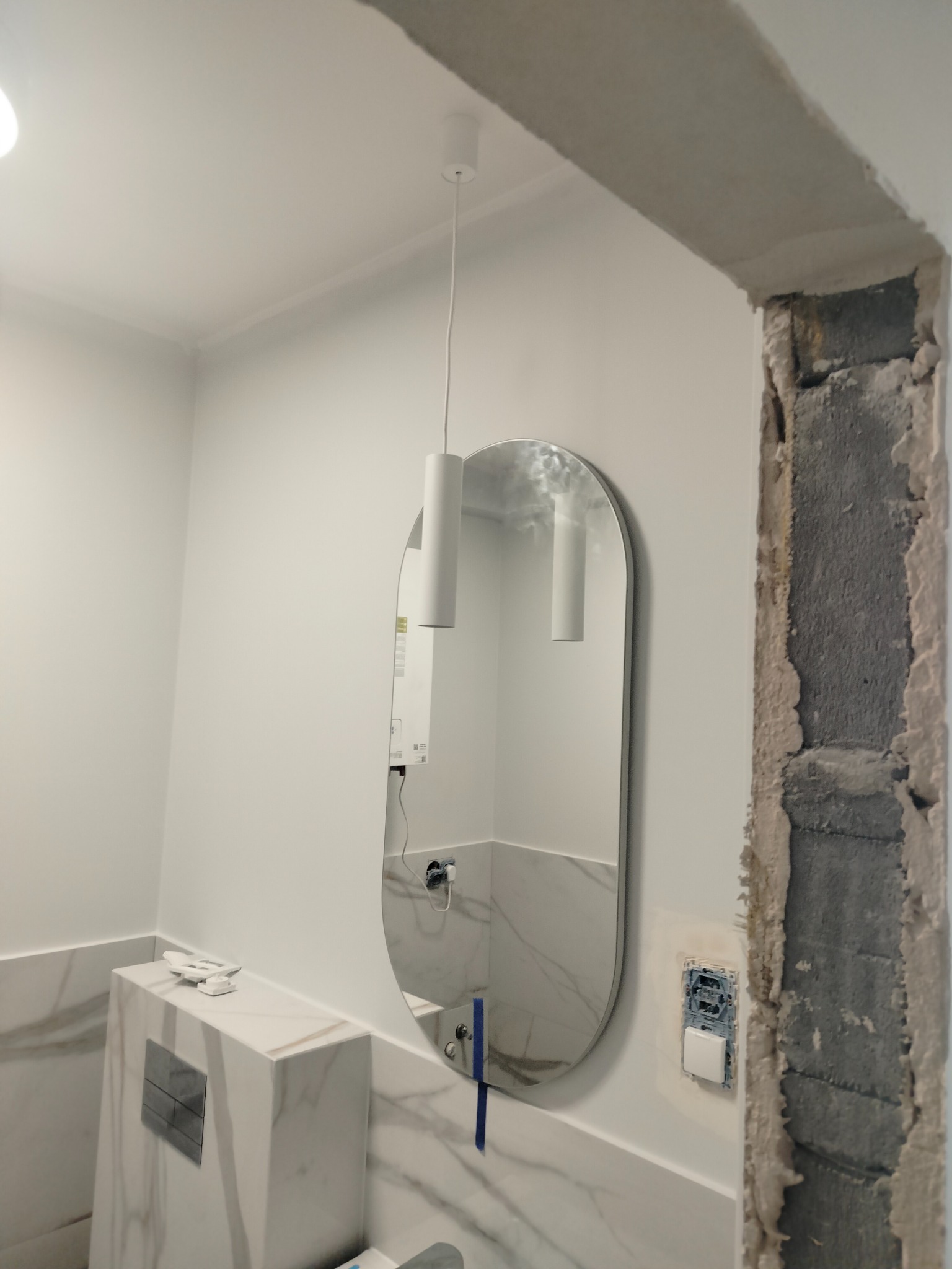 Nowoczesna łazienka w trakcie wykańczania: owalne lustro, wisząca lampa, zabudowa toalety z marmurowym wzorem, surowy fragment ściany z odsłoniętym betonem obok włącznika światła.