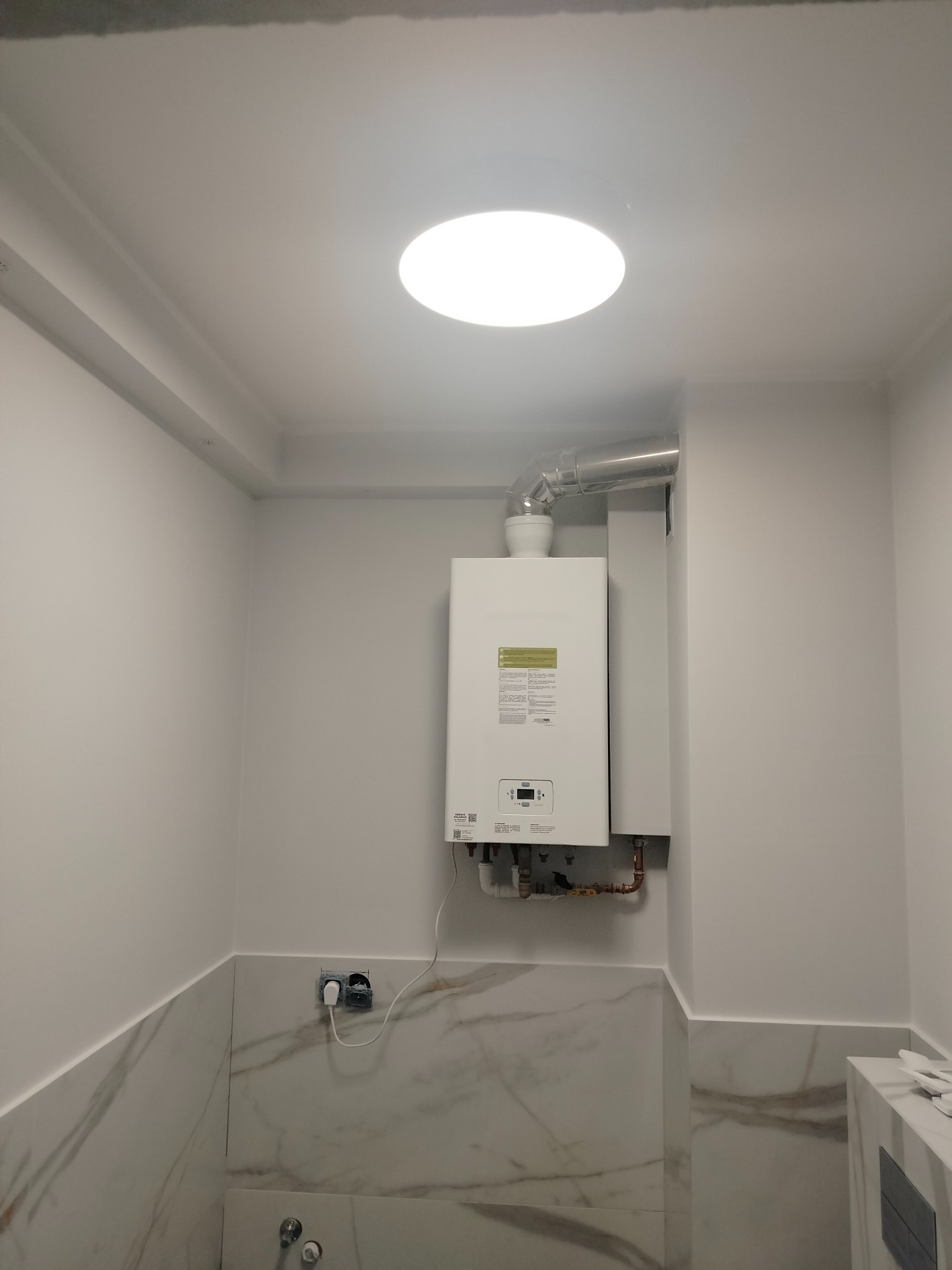 Biały, nowoczesny piec gazowy z metalową rurą odprowadzającą spaliny zamontowany na ścianie wyłożonej marmurową płytką w łazience z oświetleniem sufitowym.