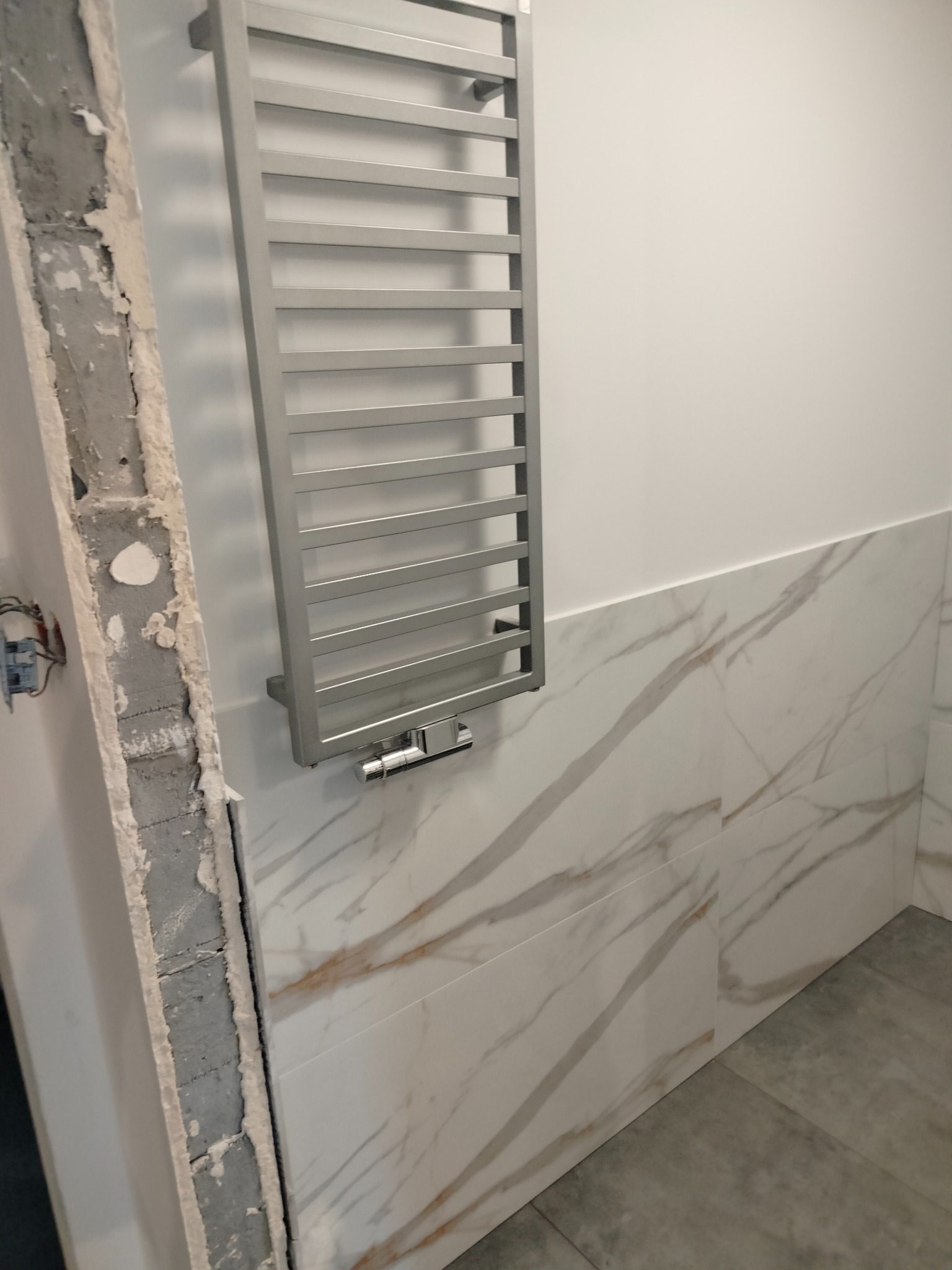 Łazienka w trakcie remontu: częściowo położone marmurowe płytki, grzejnik drabinkowy na ścianie, odsłonięta ściana z instalacją elektryczną.