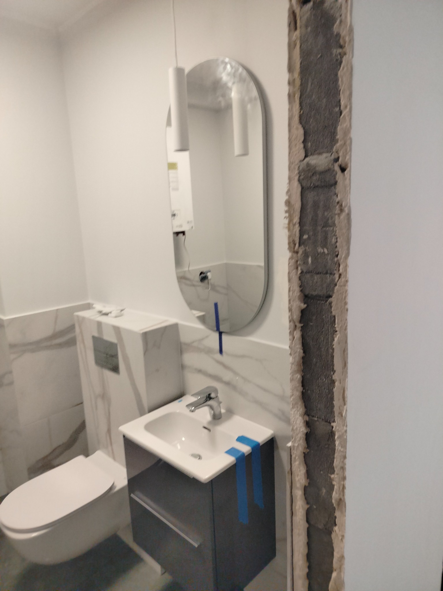 Nowoczesna łazienka w trakcie wykańczania: podwieszana toaleta, umywalka z szafką i owalne lustro, widoczne ślady prac murarskich przy ścianie.
