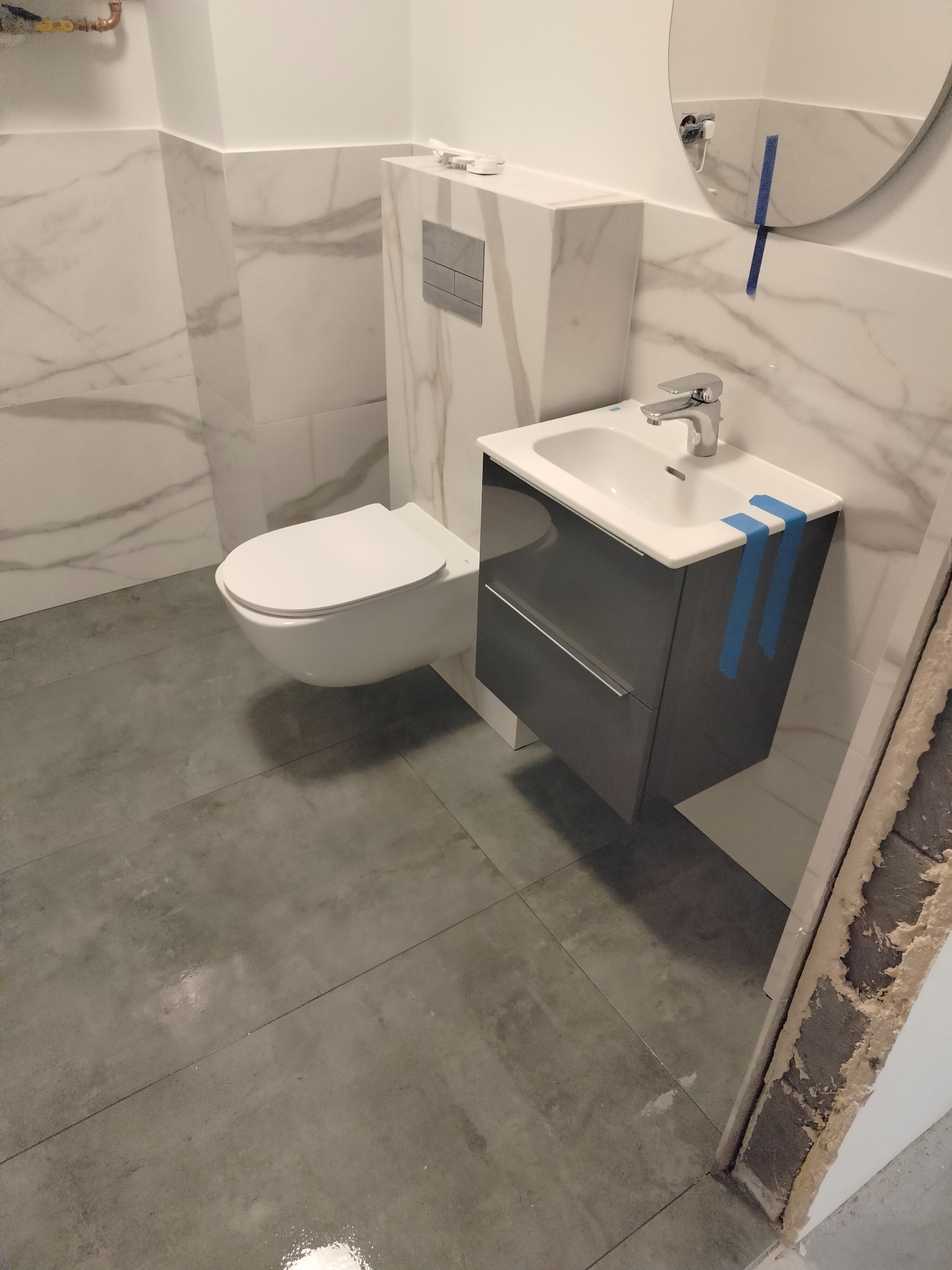 Nowoczesna łazienka z podwieszaną toaletą, szarą szafką pod umywalką i marmurowymi płytkami na ścianach, zabezpieczonymi niebieską taśmą malarską.