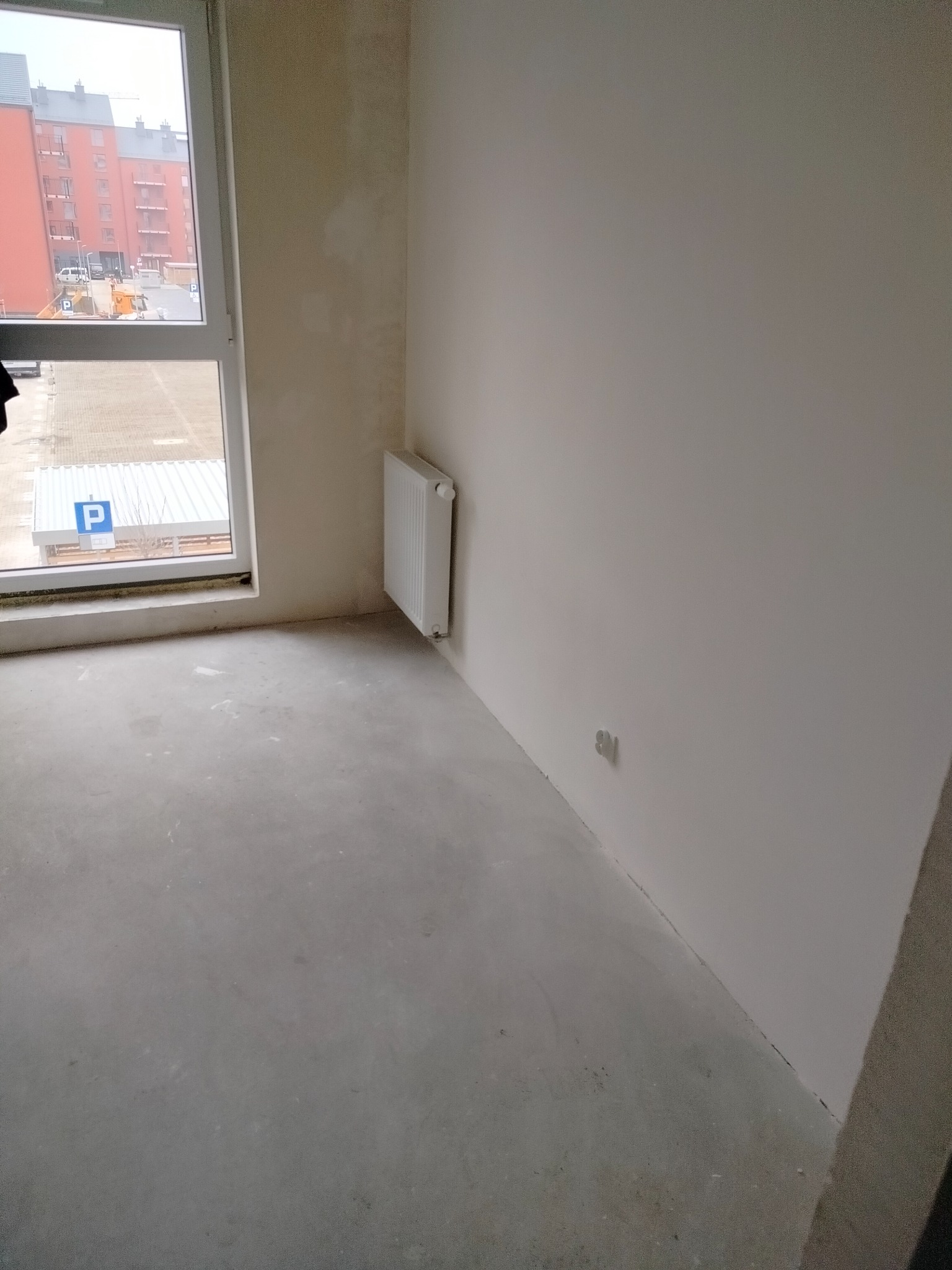 Surowe wnętrze pokoju w stanie deweloperskim: gładkie, szare wylewki podłogowe, świeżo pomalowane na biało ściany, grzejnik panelowy zamontowany pod oknem, widok na osiedle mieszkaniowe za oknem.