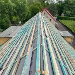 Widok z góry na konstrukcję dachu z hydroizolacją i łatami, przygotowaną do położenia pokrycia dachowego; w tle budynki mieszkalne i zieleń.