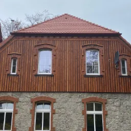 Fasada dwukondygnacyjnego domu z kamienia i drewna, z nową czerwoną dachówką, widoczne okna z ceglanymi obramowaniami i anteną satelitarną.