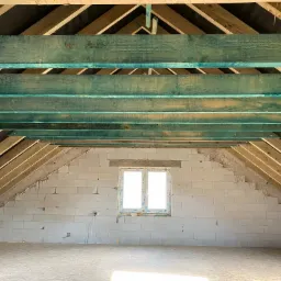 Poddasze w trakcie adaptacji: surowe ściany z bloczków, drewniana konstrukcja dachu z belek pomalowanych na turkusowo, okno w szczytowej ścianie.