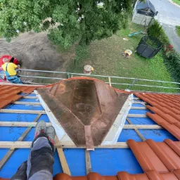 Montaż miedzianej obróbki blacharskiej na dachu krytym czerwoną dachówką ceramiczną, widok z góry na częściowo ukończony fragment dachu z widocznymi elementami konstrukcji drewnianej...