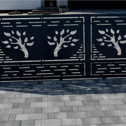 Łukasz Binkowski - Nowoczesna, czarna brama wjazdowa z wycinanymi motywami drzew, ustawiona na szarej kostce brukowej przed domem.