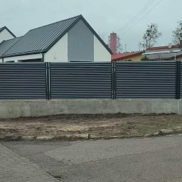 Łukasz Binkowski - Długie, nowoczesne ogrodzenie z poziomych, szarych paneli zamontowane na betonowej podmurówce, otaczające posesję z domem o szarym dachu, widoczne z poziomu ulicy.