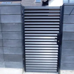 Nowoczesna brama wjazdowa z poziomymi lamelami, wkomponowana w mur z betonowych bloczków w odcieniach szarości, na tle domu z panelami słonecznymi.