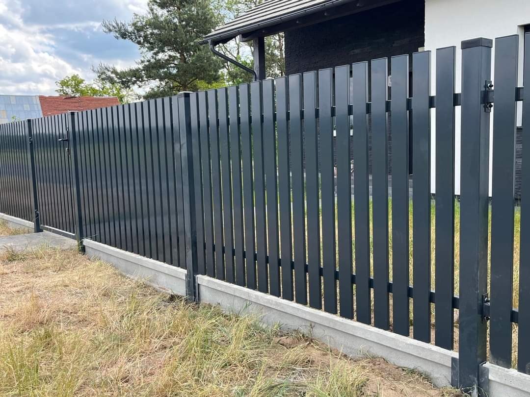 Nowoczesne, pionowe ogrodzenie z metalowych sztachet w kolorze antracytowym, osadzone na betonowej podmurówce, z furtką po lewej stronie, widoczne trawy na pierwszym planie.