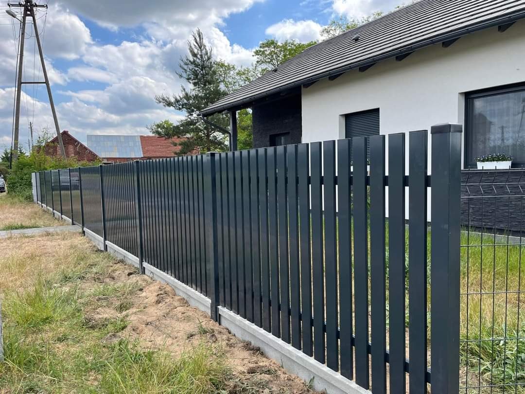 Nowoczesne, pionowe ogrodzenie panelowe w kolorze grafitowym z betonową podmurówką, na tle domu z ciemną dachówką i błękitnego nieba z chmurami.