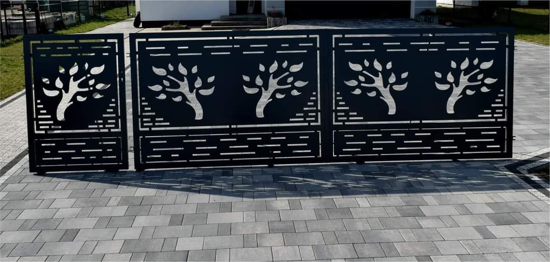 Nowoczesna, czarna brama wjazdowa z wycinanymi motywami drzew, ustawiona na szarej kostce brukowej przed domem.