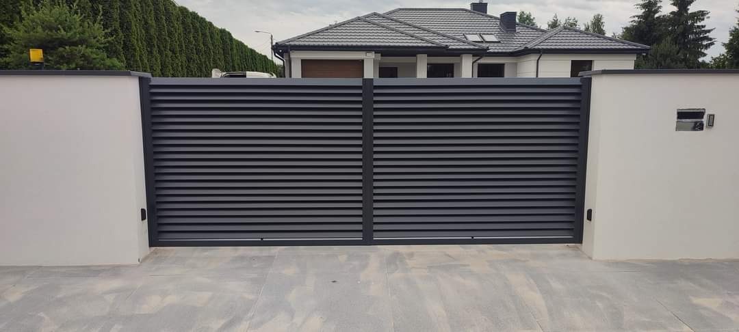 Nowoczesna, pozioma brama wjazdowa z szarych paneli, zamontowana między dwoma białymi słupkami, na tle domu z ciemnym dachem i zadbanym żywopłotem.