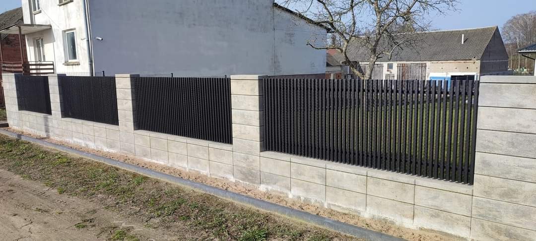 Nowoczesne ogrodzenie z betonowych bloczków i pionowych, metalowych sztachet w kolorze grafitowym, z widoczną zabudową w tle.
