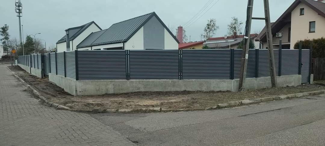 Długie, nowoczesne ogrodzenie z poziomych, szarych paneli zamontowane na betonowej podmurówce, otaczające posesję z domem o szarym dachu, widoczne z poziomu ulicy.