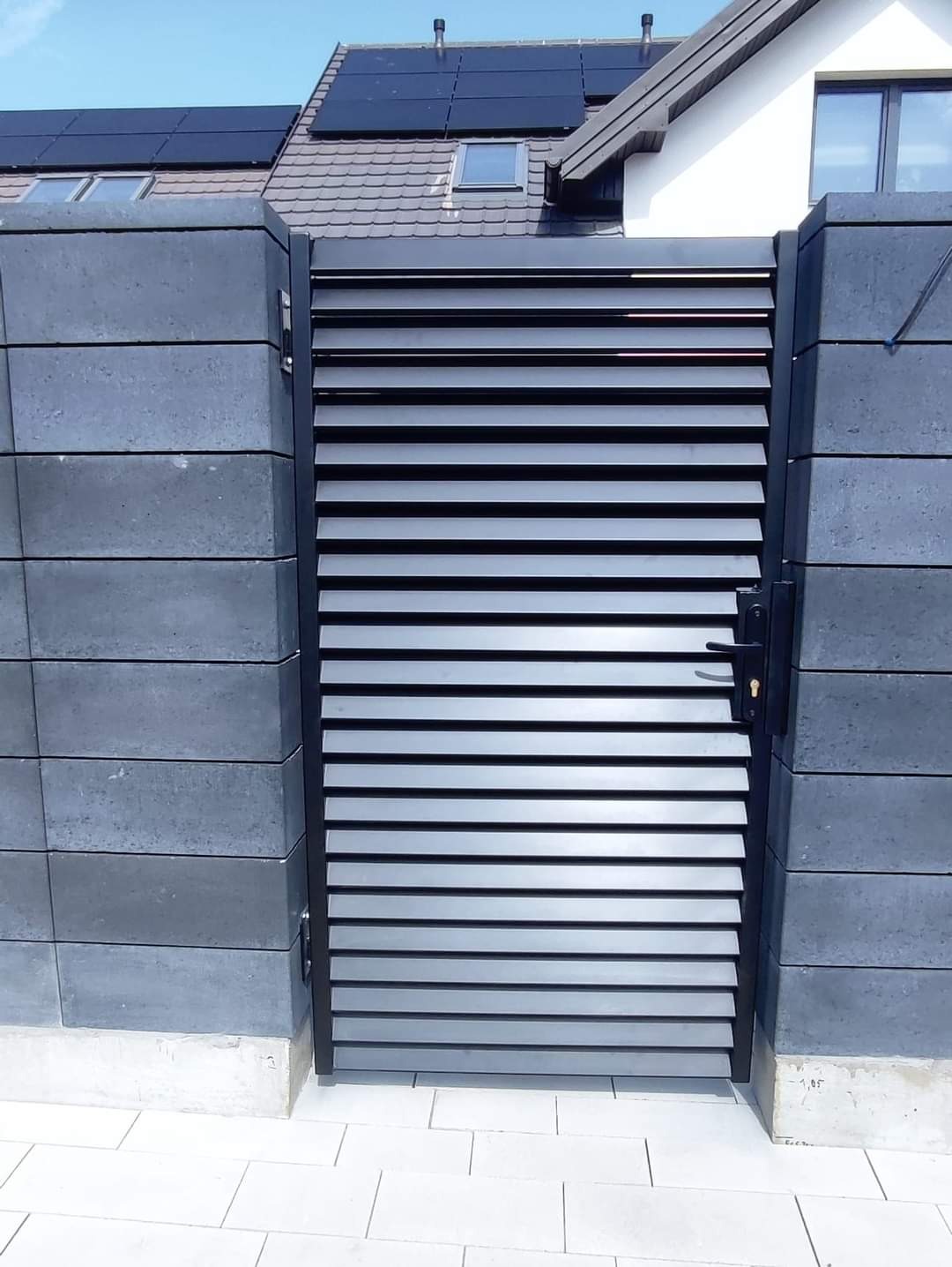 Nowoczesna brama wjazdowa z poziomymi lamelami, wkomponowana w mur z betonowych bloczków w odcieniach szarości, na tle domu z panelami słonecznymi.