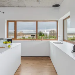 Nowoczesna, minimalistyczna kuchnia w bieli z drewnianymi oknami, drewnianą podłogą i widokiem na jezioro, z dekoracjami: teleskop, owoce i deski do krojenia.