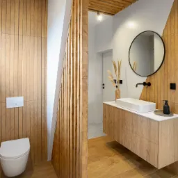 Nowoczesna łazienka z drewnianymi panelami ściennymi i lamelami, wisząca szafka z umywalką nablatową, okrągłe lustro w czarnej ramie i drewniany sufit z oświetleniem punktowym.