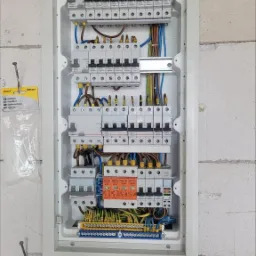 Rozdzielnica elektryczna z zabezpieczeniami różnicowoprądowymi i nadprądowymi, widoczne przewody, listwy zaciskowe, zamocowana na ścianie z bloczków, obok opakowanie opasek zaciskowych Anslut.