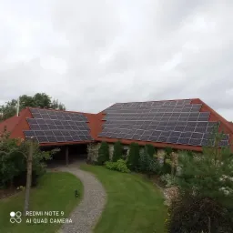 Dom z czerwoną dachówką i panelami słonecznymi na dachu, widok z góry na posesję z kamienną ścieżką i zadbanym trawnikiem w pochmurny dzień.