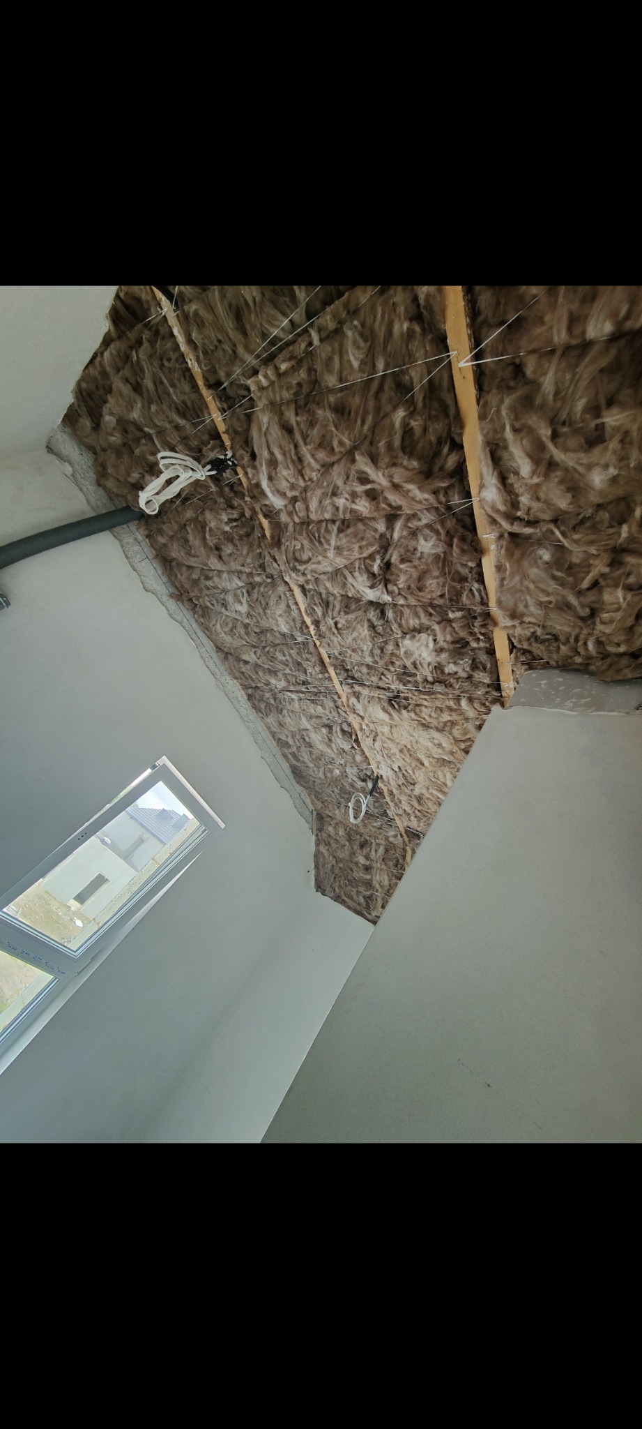 Izolacja poddasza wełną mineralną, widoczne drewniane belki konstrukcyjne i przewody elektryczne, surowe ściany i okno dachowe w nowym domu.