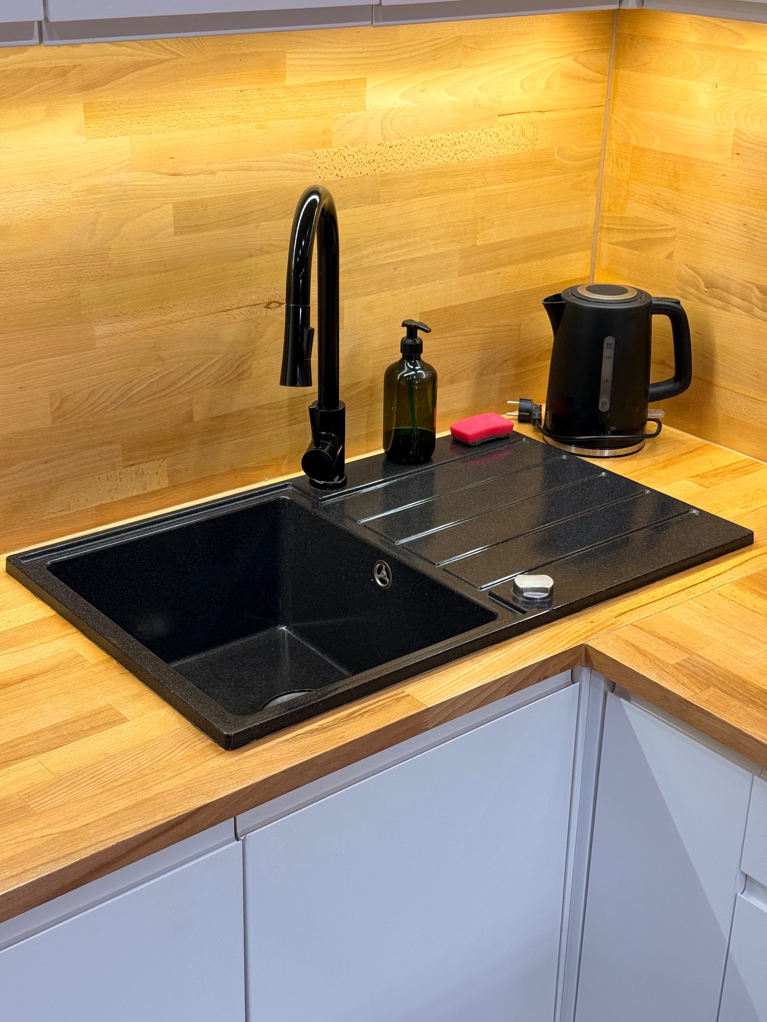 Wypolerowany, czarny, granitowy zlew kuchenny z baterią, dozownikiem mydła i czajnikiem na blacie z jasnego drewna, po sprzątaniu. Widoczne białe szafki.