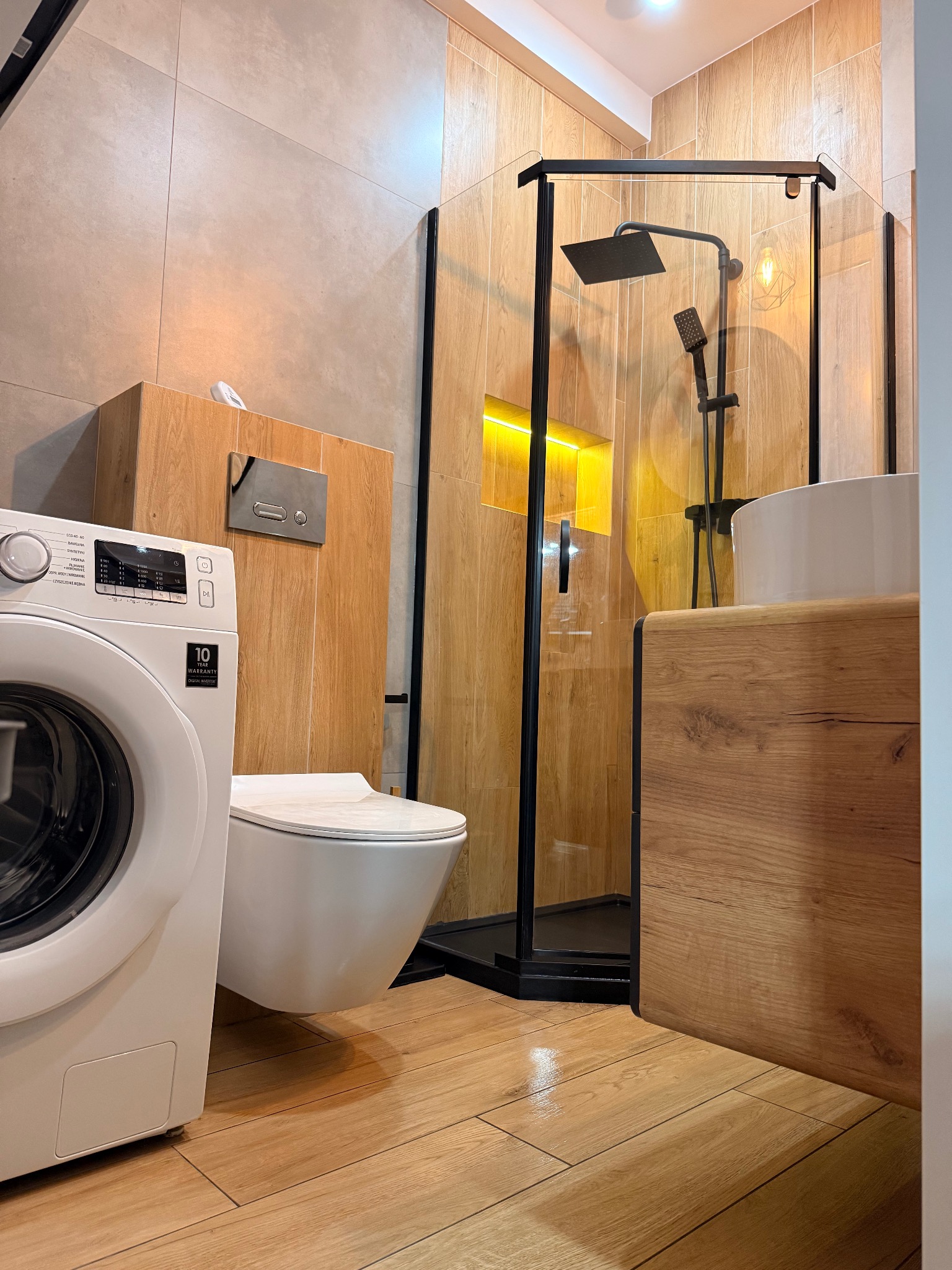 Nowoczesna łazienka po remoncie: kabina prysznicowa typu walk-in z czarnymi ramami, pralka i toaleta w jasnych barwach, drewnopodobne płytki na podłodze i ścianach.