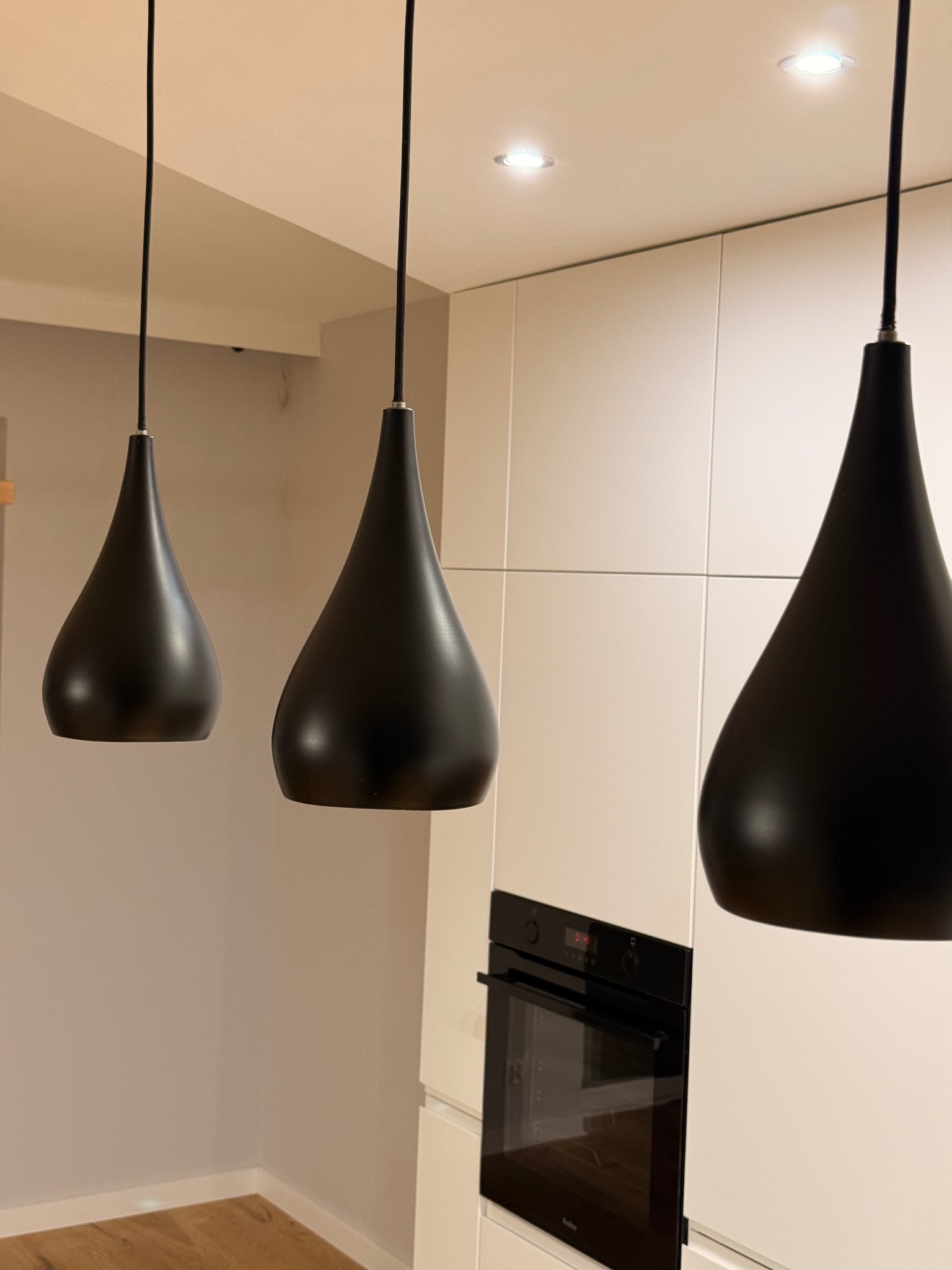 Nowoczesna kuchnia po remoncie: trzy czarne, kroplowe lampy wiszące nad białymi szafkami i piekarnikiem. Minimalistyczny design, ciepłe oświetlenie.