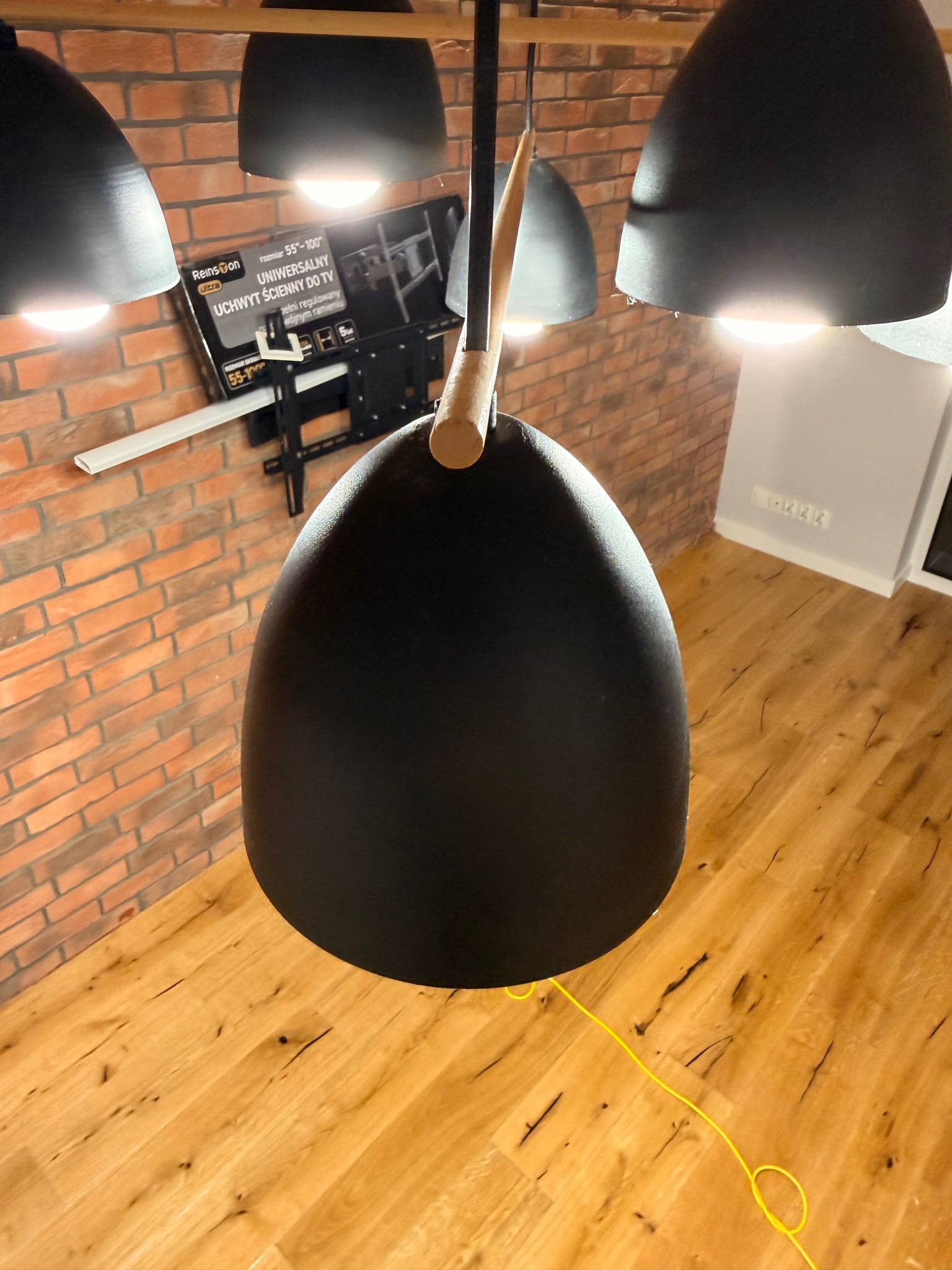 Lampa wisząca w stylu loft z ceglaną ścianą i drewnianą podłogą w tle, Warszawa. Widoczny uchwyt do TV na ścianie. Żółty kabel na podłodze dodaje koloru.