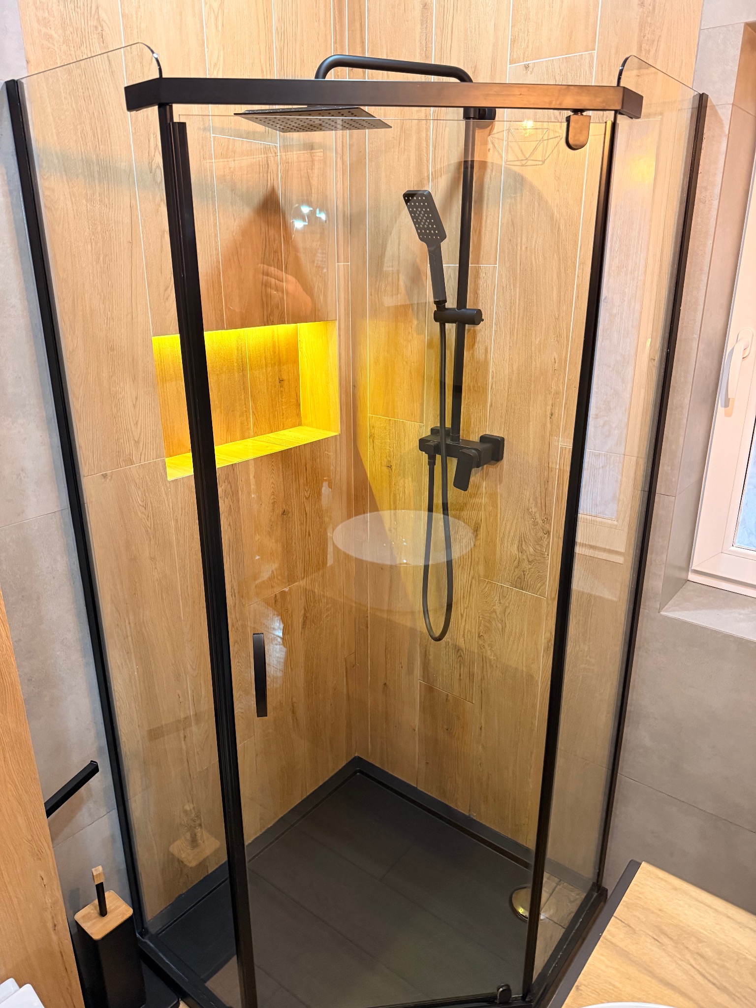 Nowoczesna kabina prysznicowa z czarnymi ramami i drewnopodobnymi płytkami, oświetlona wnęka. Widok z góry, ukazujący czystość i detale wykończenia.
