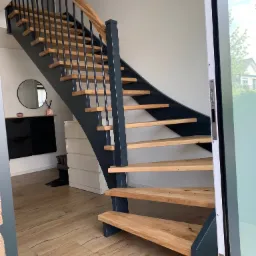 Nowoczesne schody z drewnianymi stopniami i ciemnoszarą konstrukcją, widoczne z boku, z poręczą z naturalnego drewna i czarnymi, pionowymi elementami, na tle jasnego wnętrza z drewnianą podłogą...