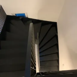 Widok z góry na czarne, spiralne schody z metalową balustradą, zabezpieczone taśmą malarską na drewnianej podłodze podczas prac wykończeniowych.