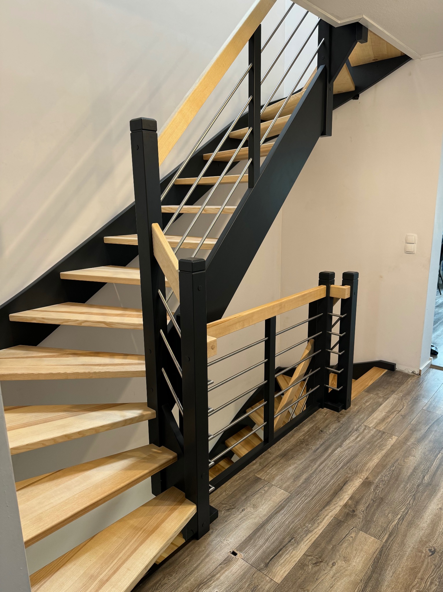 Nowoczesne schody z drewnianymi stopniami i czarną konstrukcją, z metalowymi, poziomymi prętami balustrady, na podłodze panele imitujące drewno.