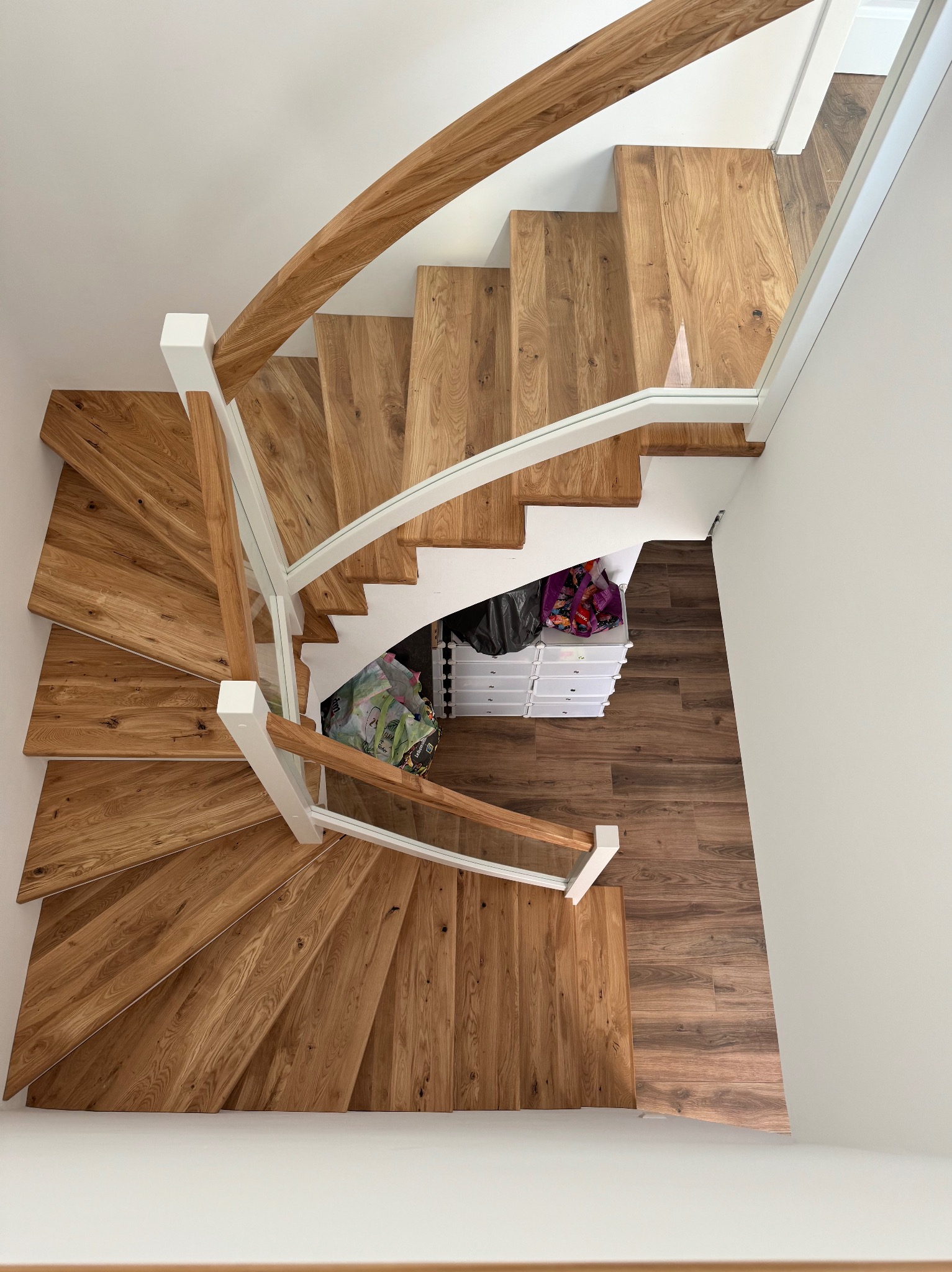 Kątowe ujęcie z góry na drewniane schody z jasnymi, drewnianymi poręczami i szklanymi panelami, prowadzące w dół do pomieszczenia z brązową podłogą i białymi szafkami z szufladami, częściowo...