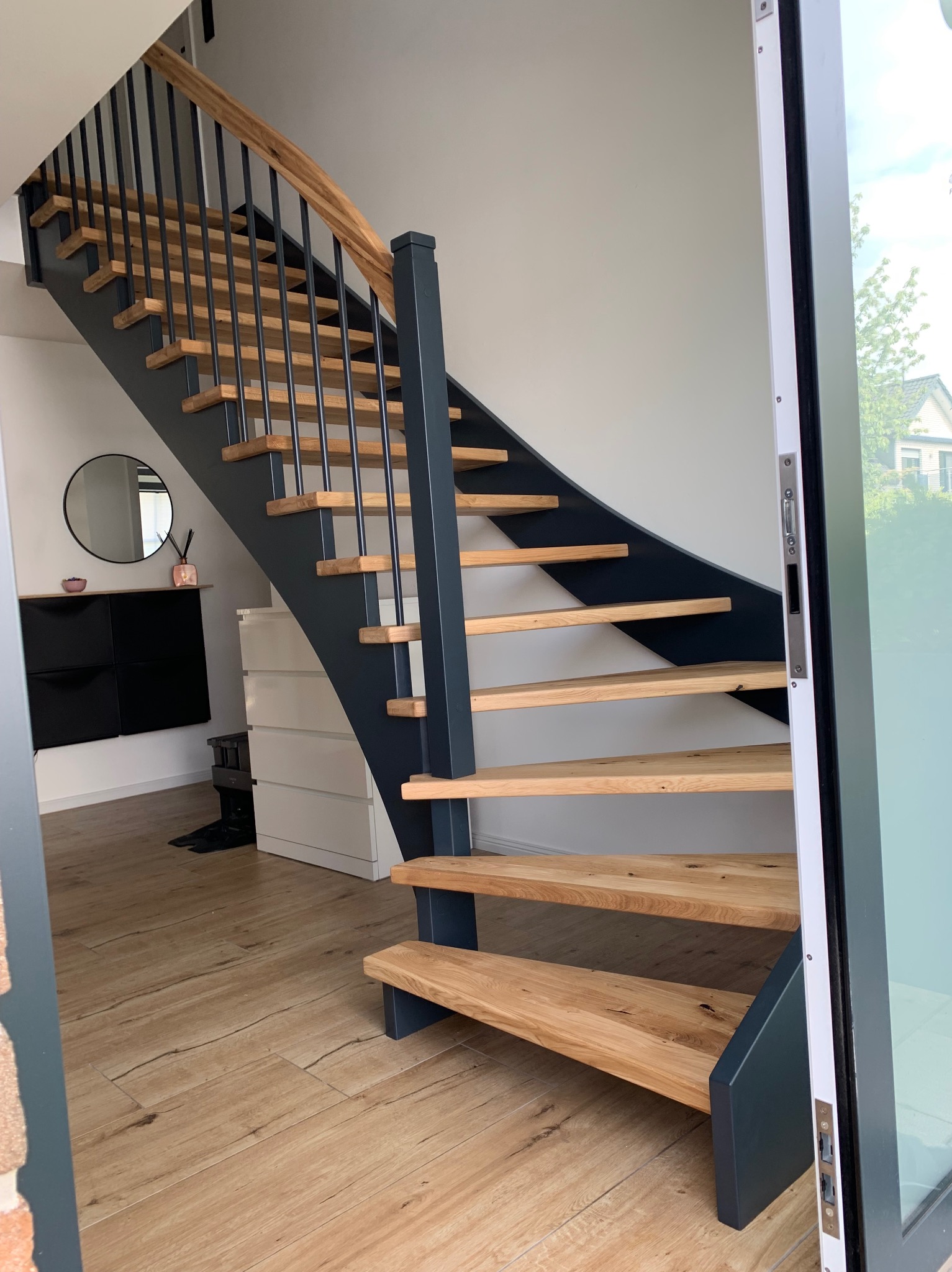 Nowoczesne schody z drewnianymi stopniami i ciemnoszarą konstrukcją, widoczne z boku, z poręczą z naturalnego drewna i czarnymi, pionowymi elementami, na tle jasnego wnętrza z drewnianą podłogą...