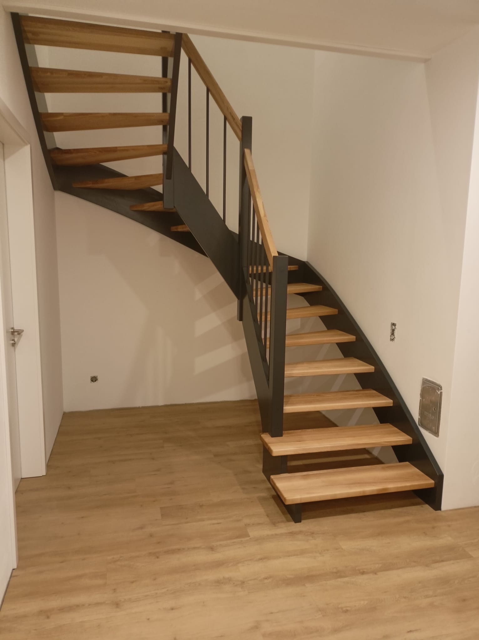 Nowoczesne schody z drewnianymi stopniami i metalową konstrukcją, widoczne od dołu, z drewnianą poręczą, w jasnym wnętrzu z panelami podłogowymi.