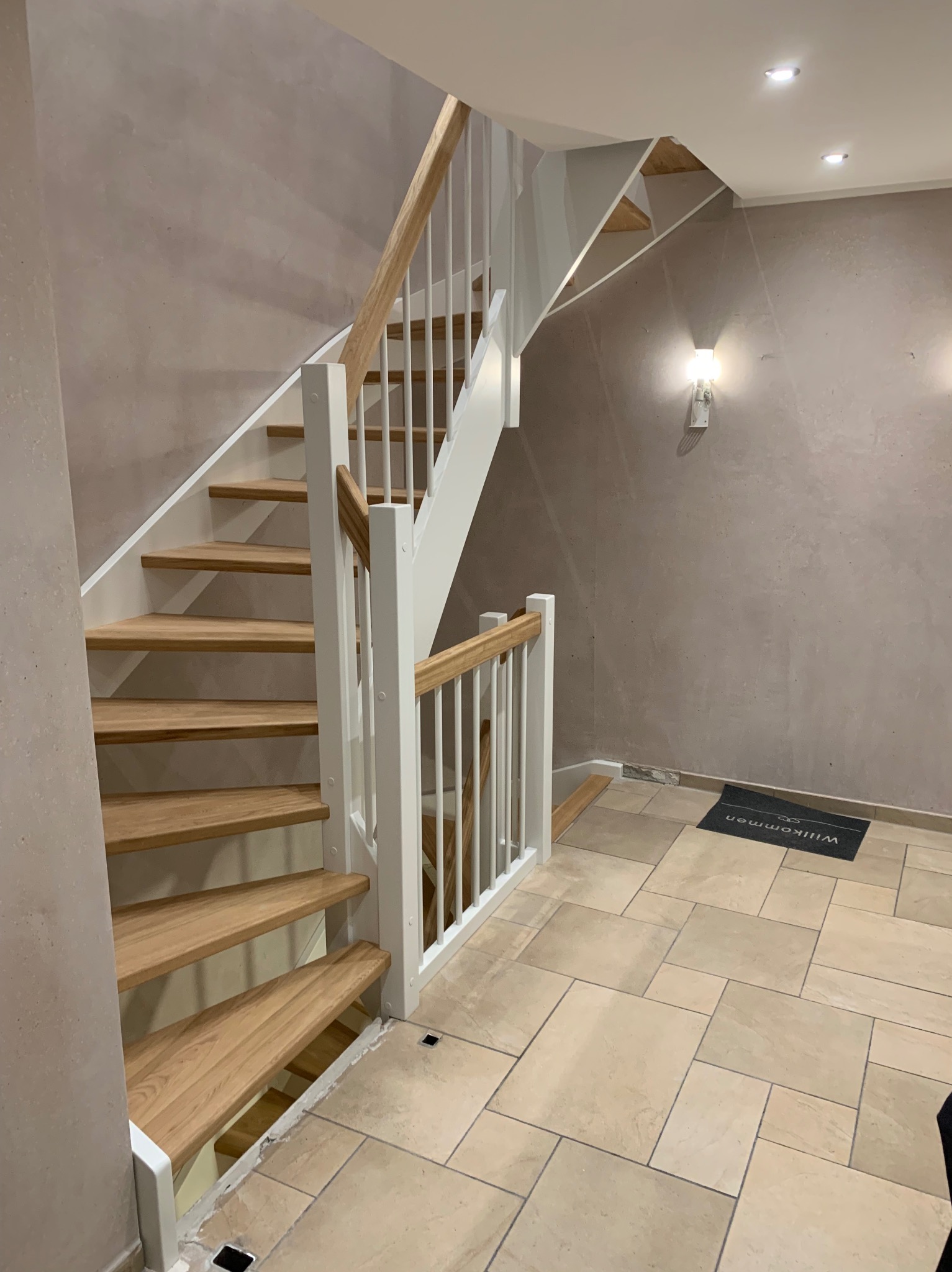 Drewniane schody z białymi balustradami w nowoczesnym wnętrzu, widok z dołu na pierwsze stopnie i fragment podestu, na podłodze płytki i wycieraczka.