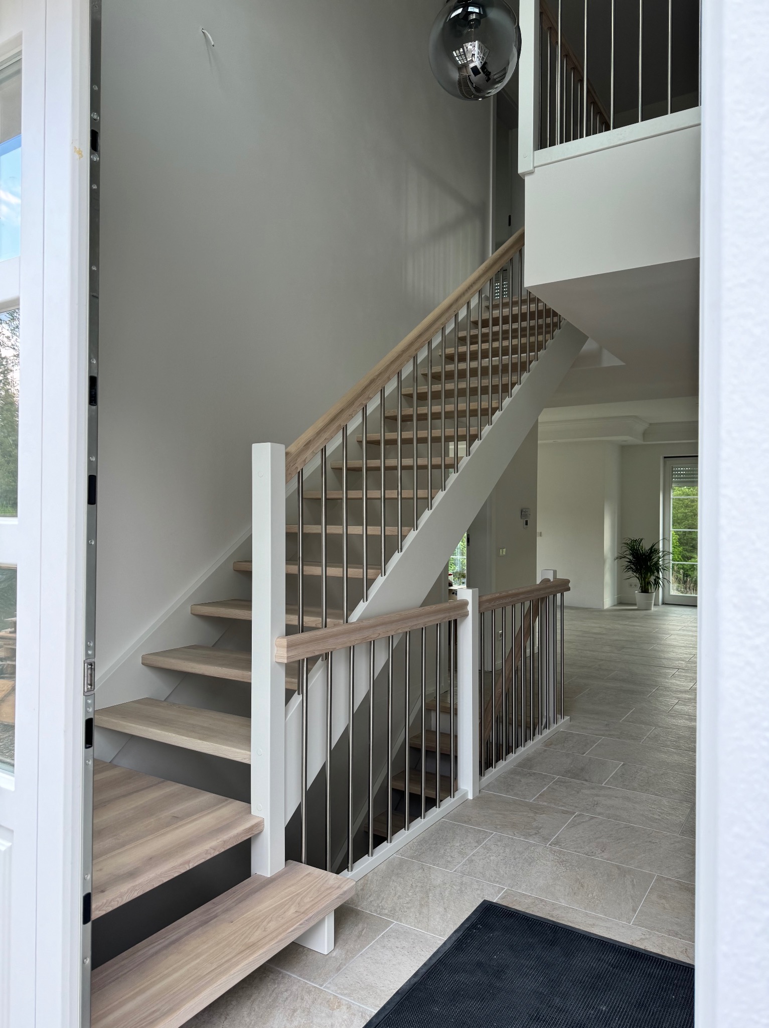 Nowoczesne schody dwubiegowe z drewnianymi stopniami i metalowymi balustradami, widoczne z perspektywy wejścia do domu, z czarną wycieraczką u dołu.