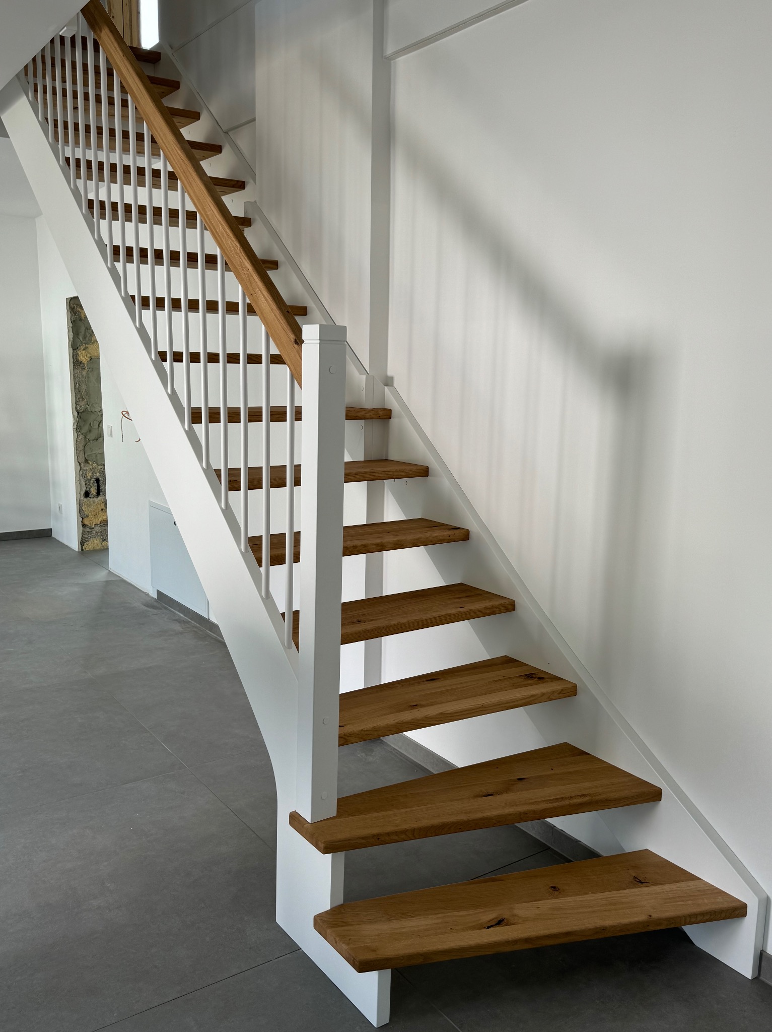 Nowoczesne schody z drewnianymi stopniami i białą balustradą, widok z dołu, szare płytki na podłodze, minimalistyczne wnętrze.