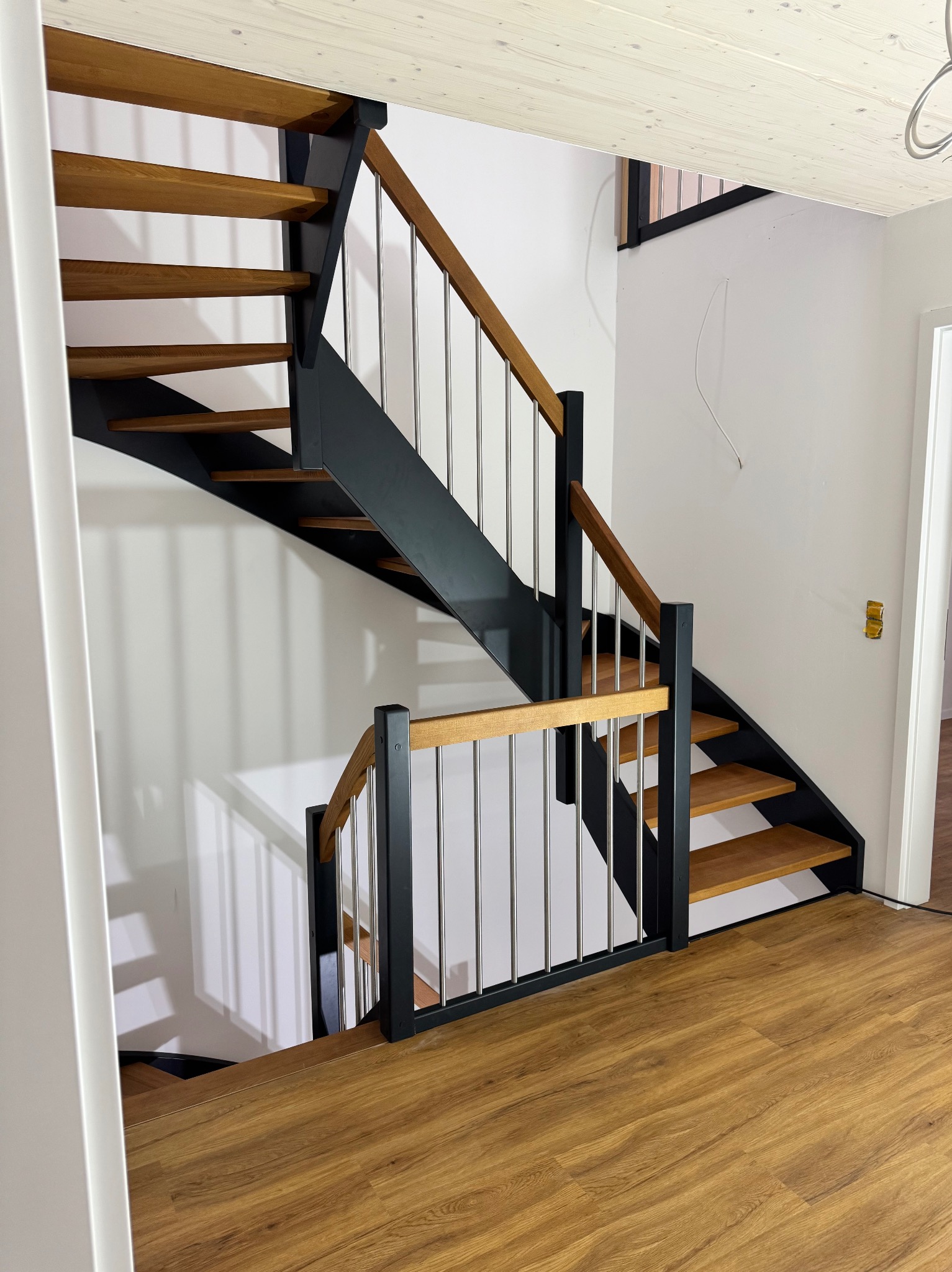 Nowoczesne schody z drewnianymi stopniami i poręczami z metalowymi prętami, czarne elementy konstrukcyjne, widok z dołu na tle białej ściany i drewnianej podłogi.